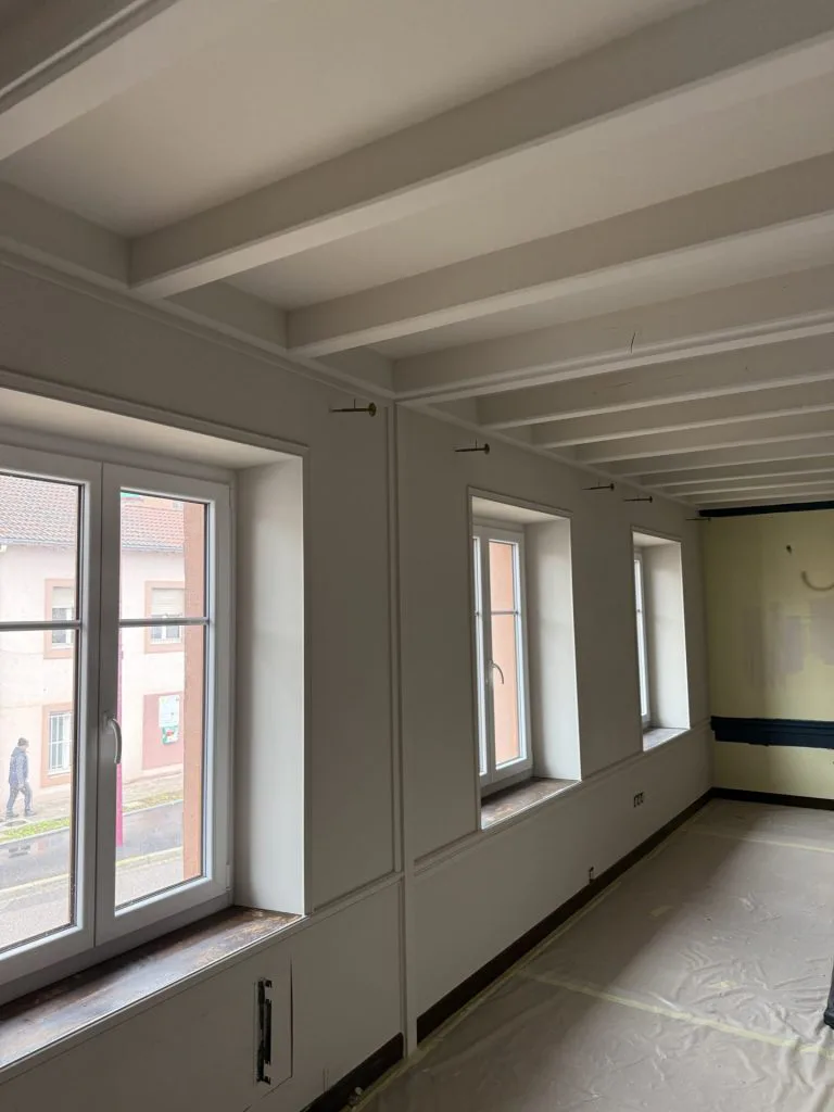 Pièce de vie en cours de rénovation avec poutres peintes en blanc et grandes fenêtres
