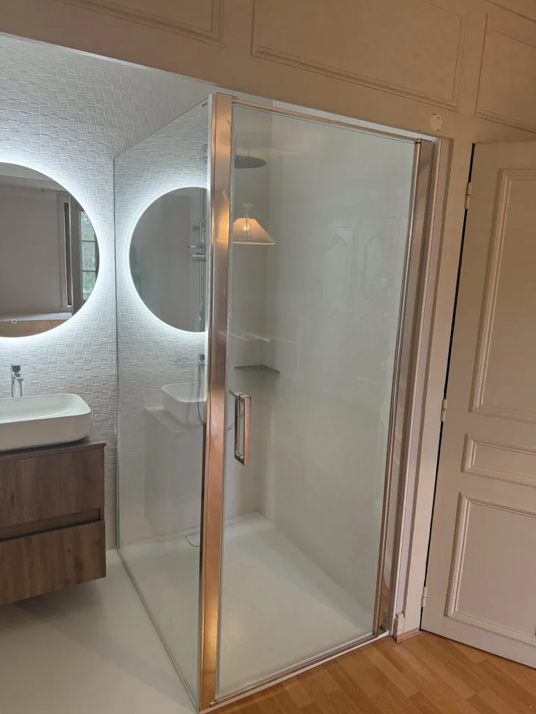 Salle d’eau vitrée intégrée à une pièce avec parquet, miroirs ronds et meuble vasque en bois