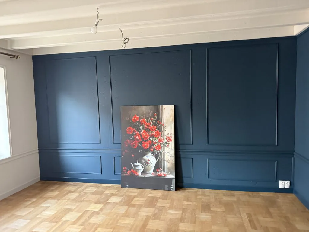 Pièce rénovée avec mur bleu à moulures, parquet et tableau floral posé au sol