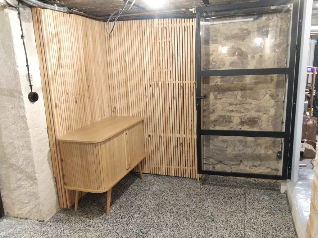 Porte vitrée noire et buffet en bois devant un mur à tasseaux de la cave rénovée