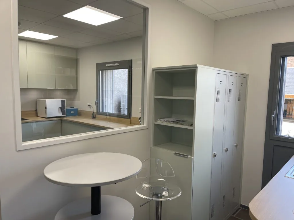 Salle du personnel avec table ronde, chaises transparentes et ouverture sur la kitchenette