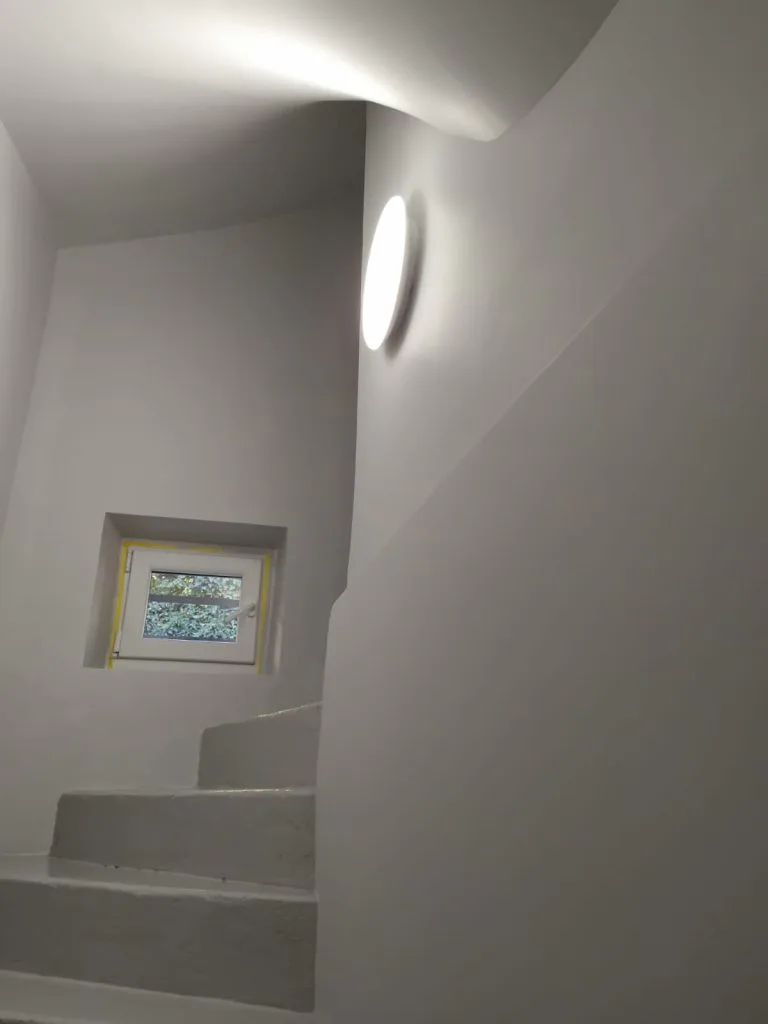 Escalier d’accès rénové avec murs blancs, applique ronde et petite fenêtre