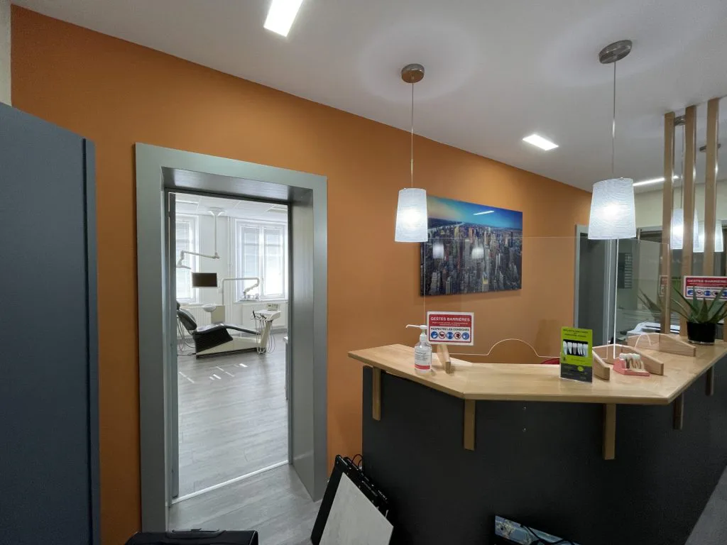 Ancien accueil du cabinet avec comptoir noir, mur orange et ouverture vers une salle de soins