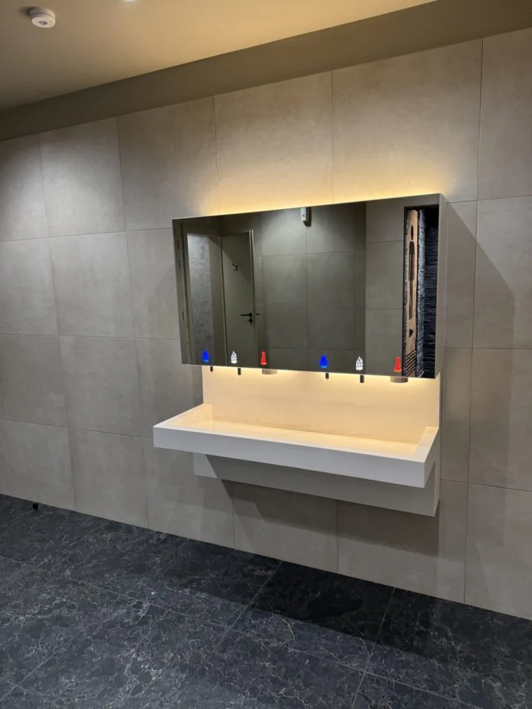 Vasque collective blanche sous un grand miroir, sur un mur de carrelage gris